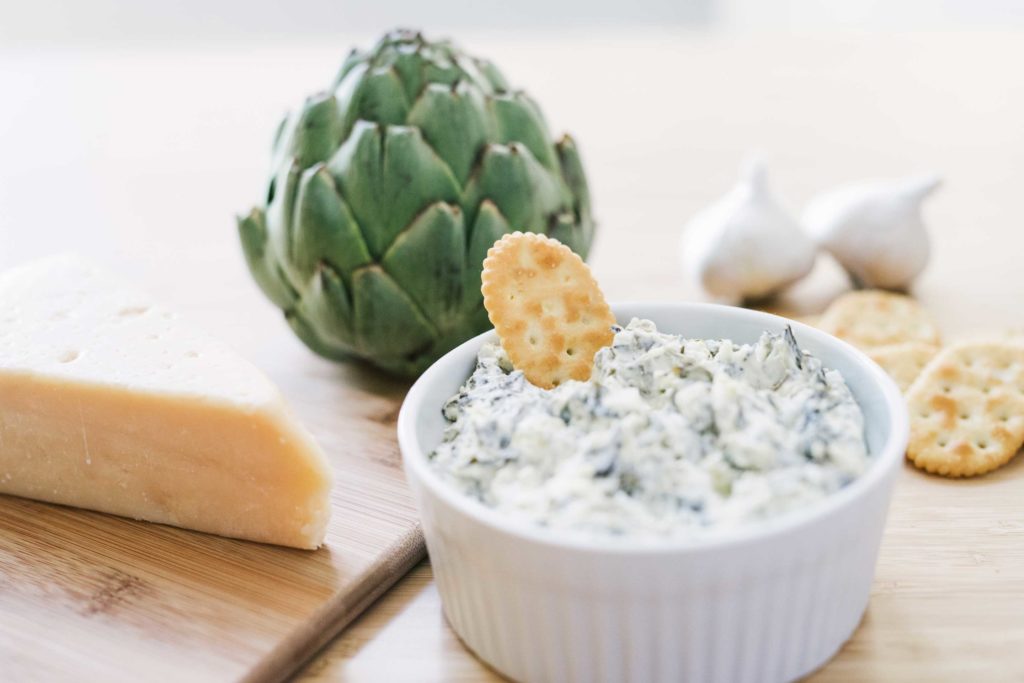 bowl of dip with a cracker in it and cheese and artichoke in the background