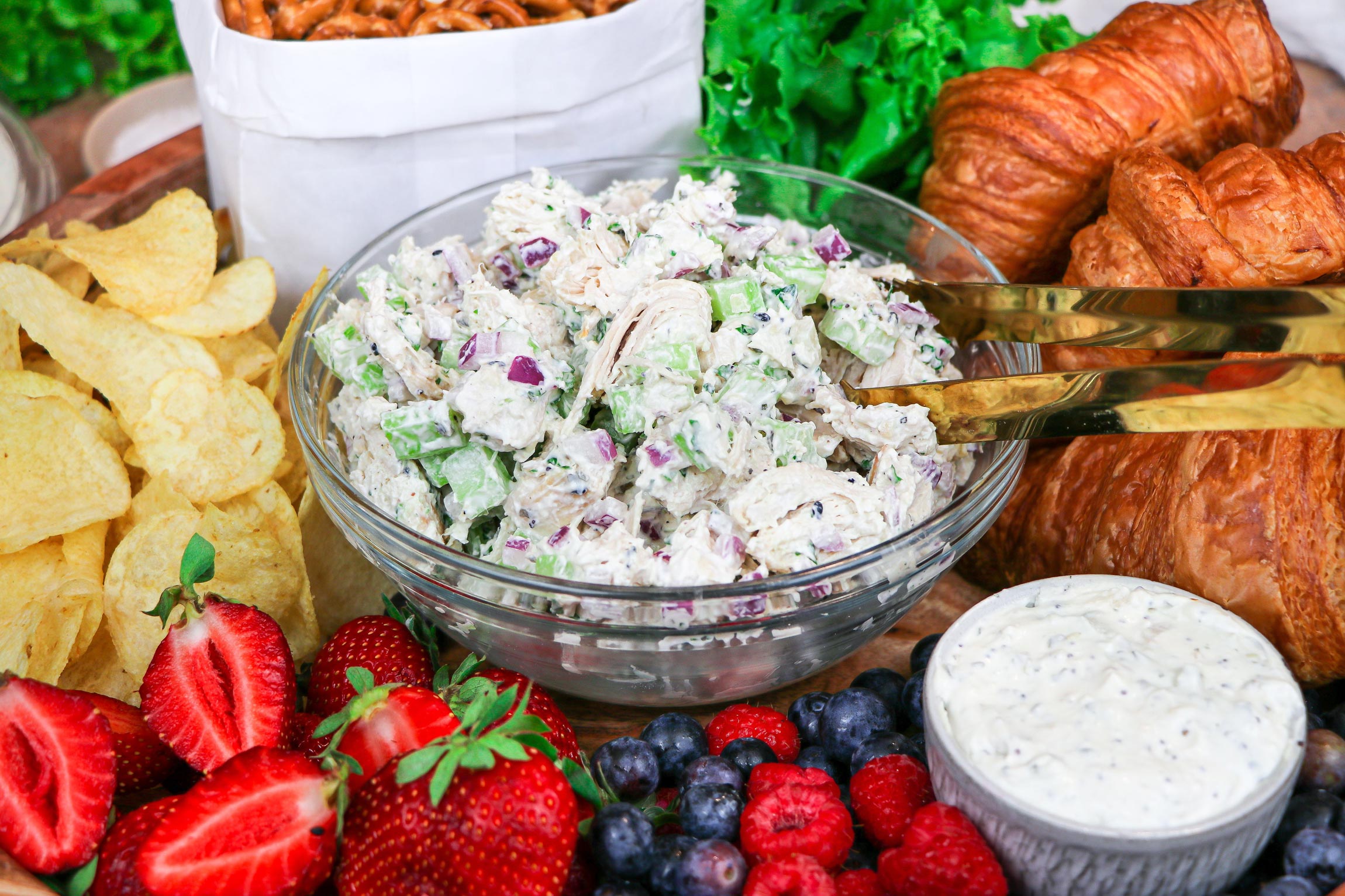 chicken salad in a bowl surrounded by fruit, chips, pretzels and dip